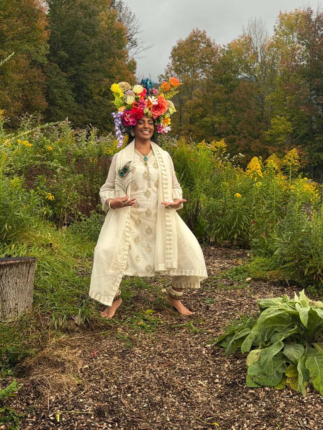 A smiling woman in white embroidery and a towering crown of coral roses, posing in a garden
