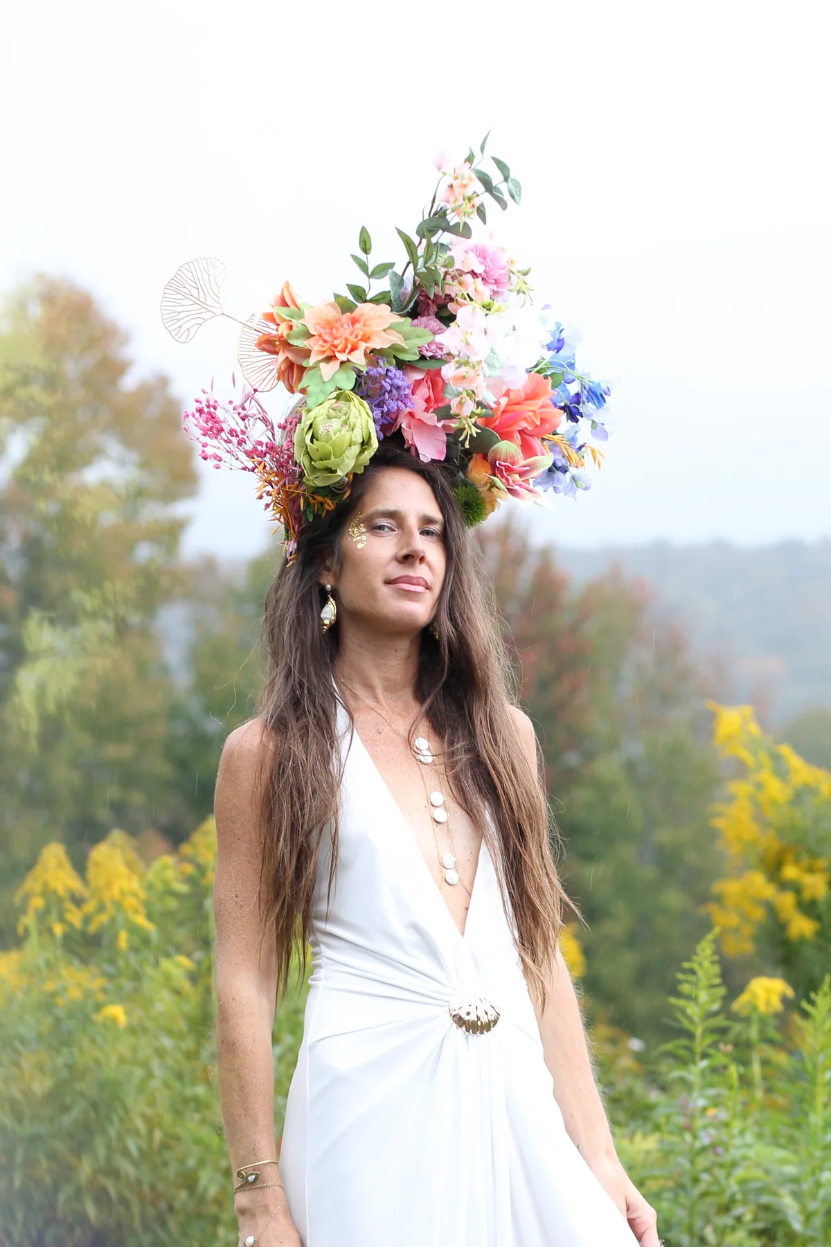 A woman in a white halter gown wearing a tall crown of peach, pink and blue flowers, photographed from the waist up in a misty garden
