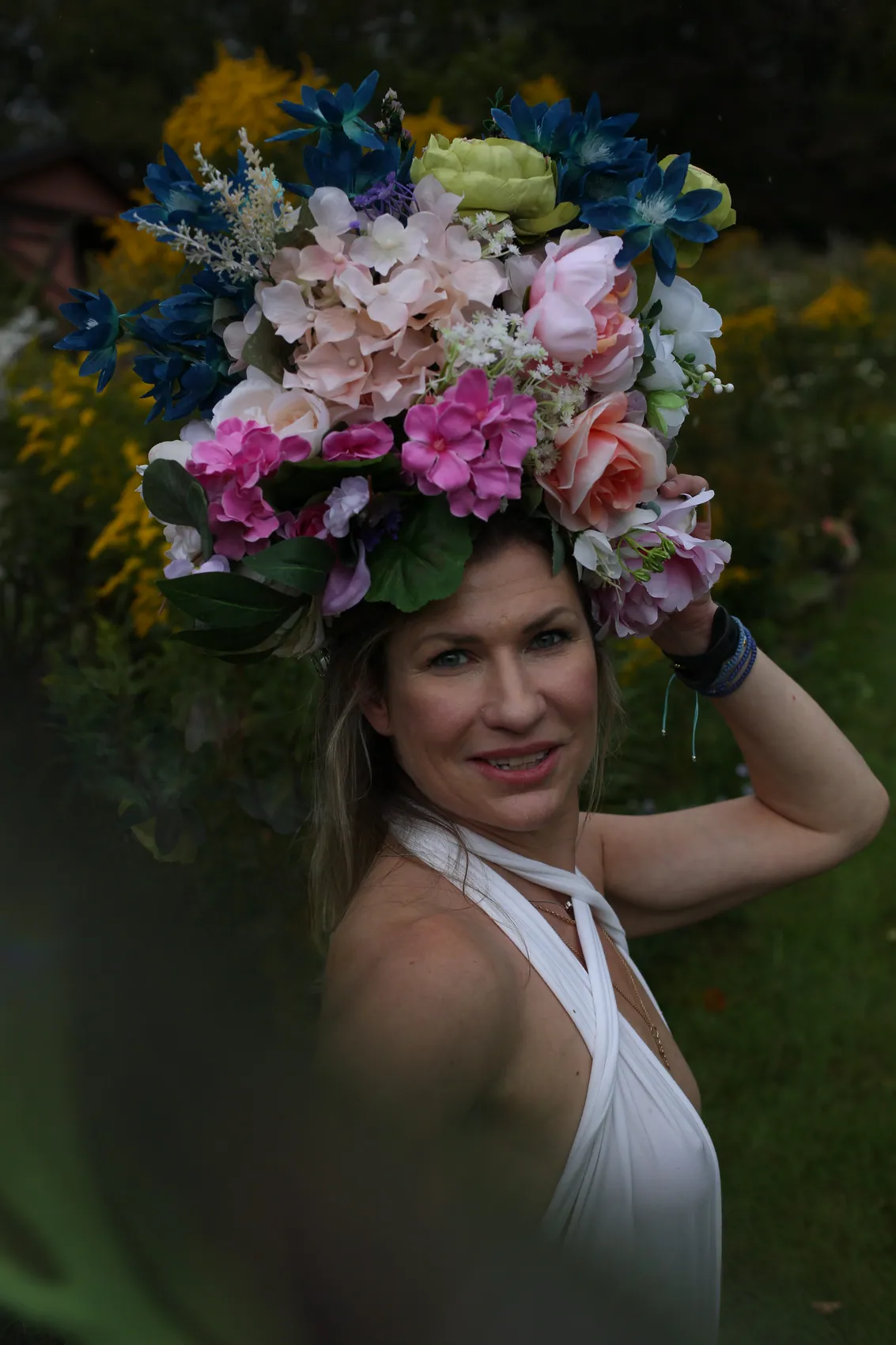 A smiling woman in a white halter top steadying a wide crown of pink hydrangea and blue lilies