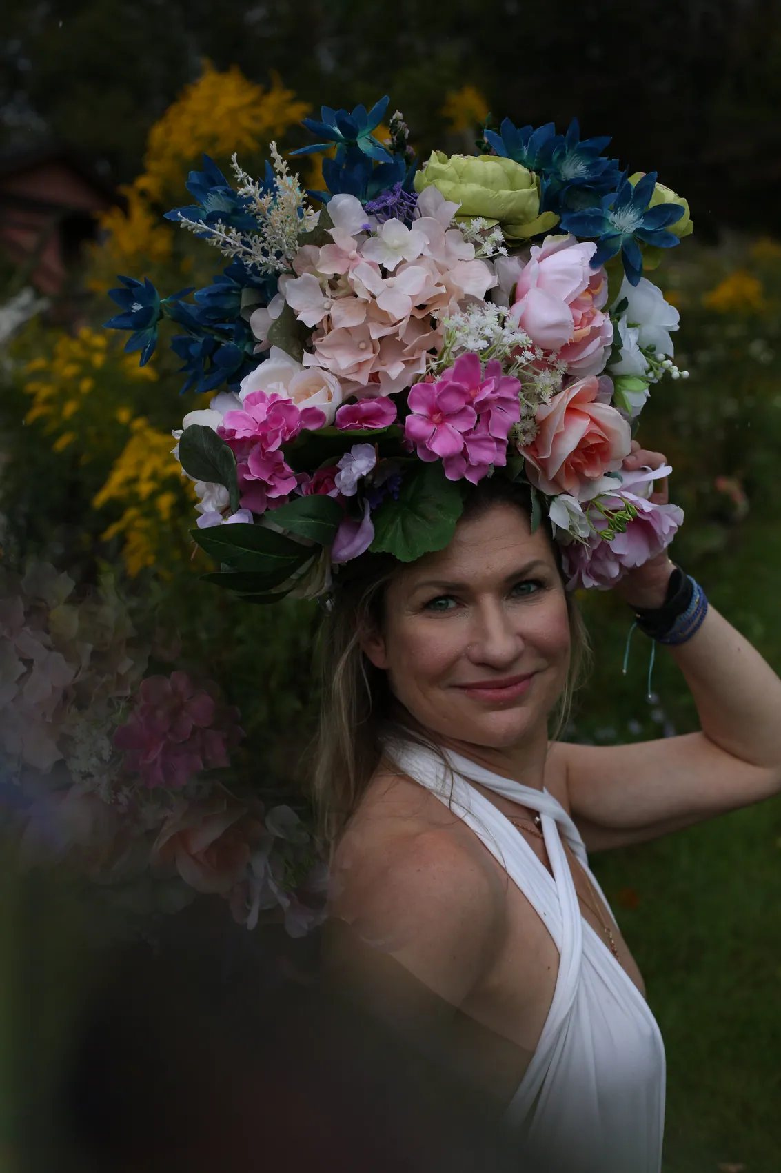 A smiling woman in a white halter top steadying a wide crown of pink hydrangea and blue lilies