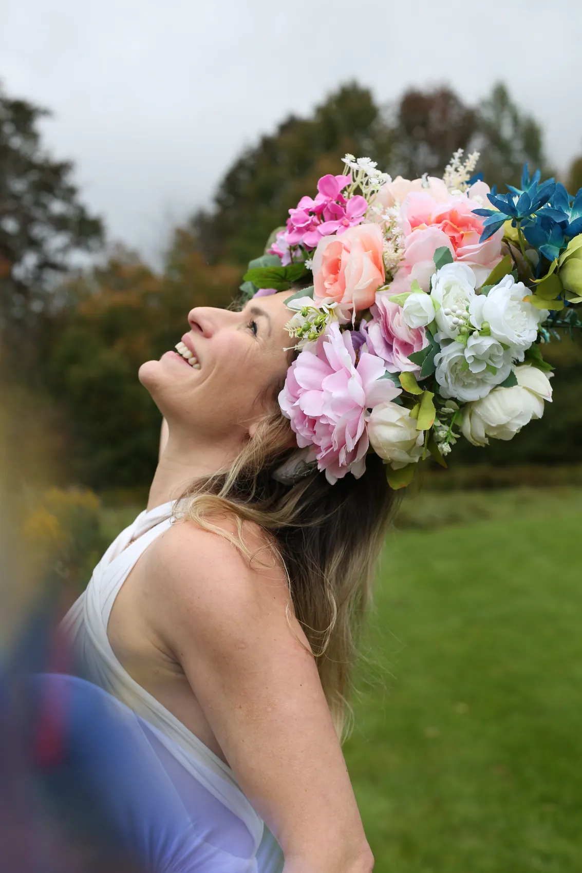 A woman in a white halter top laughing up at the sky beneath a crown of pink peonies, white roses and blue lilies