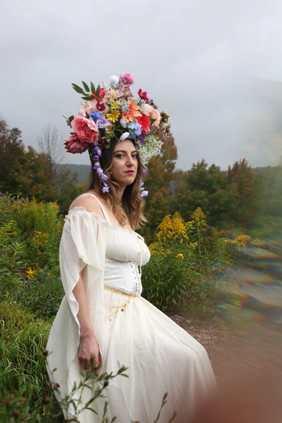 A woman in a cream off-shoulder dress wearing a tall multicolour crown with trailing wisteria