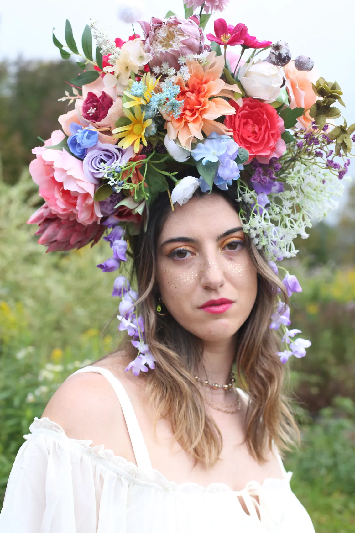 A woman with glitter freckles in an off-shoulder white blouse under a towering multicolour crown with trailing wisteria