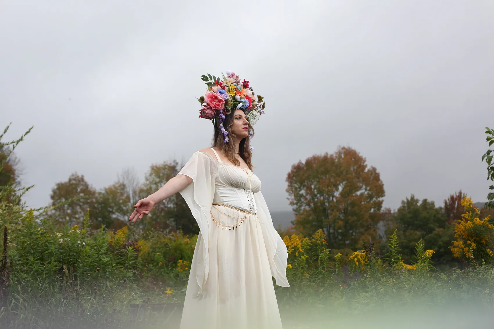 A woman in a flowing cream dress and crown with her arms outstretched under a grey sky