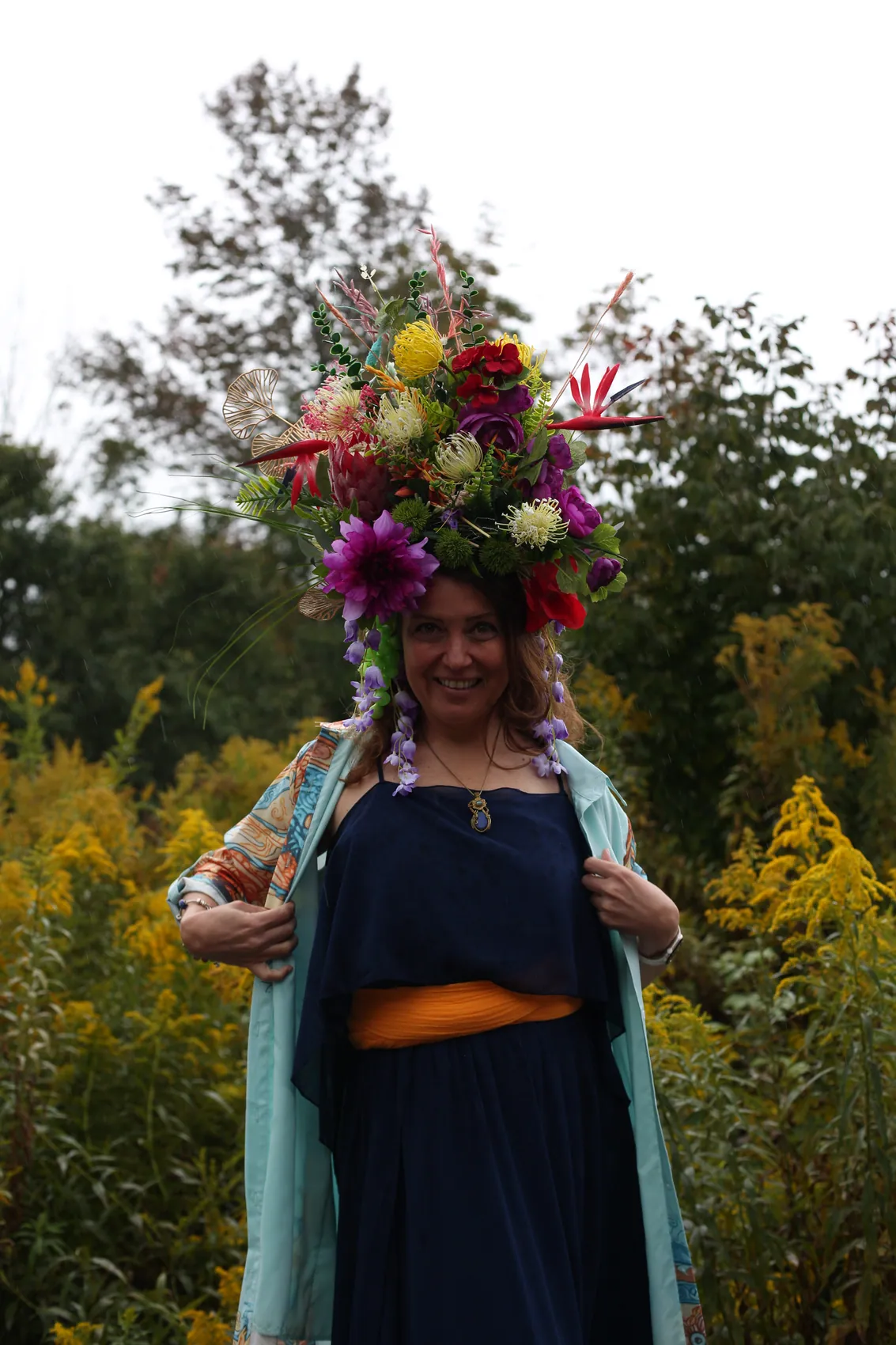 A smiling woman in a navy dress and kimono under a towering crown of dahlias