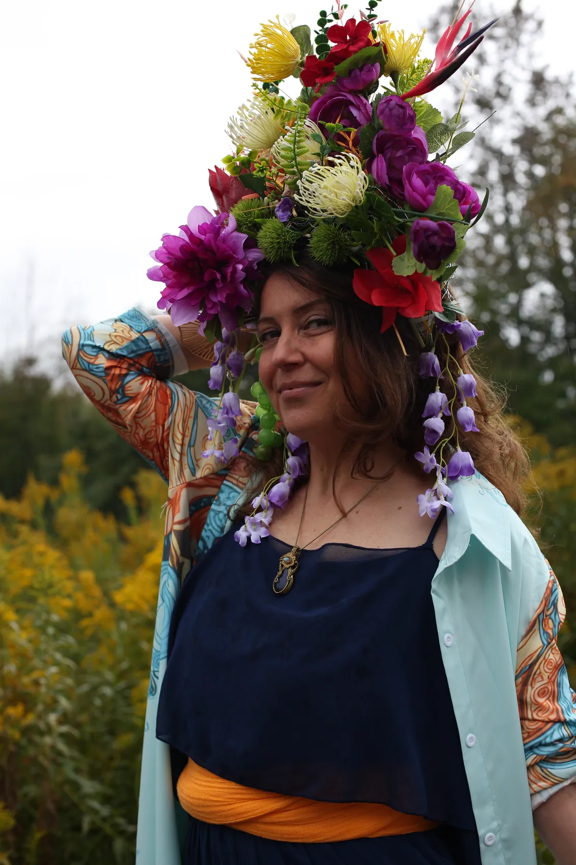 A smiling woman in a navy dress and pastel kimono, one hand in her hair, under a tall crown of dahlias and protea