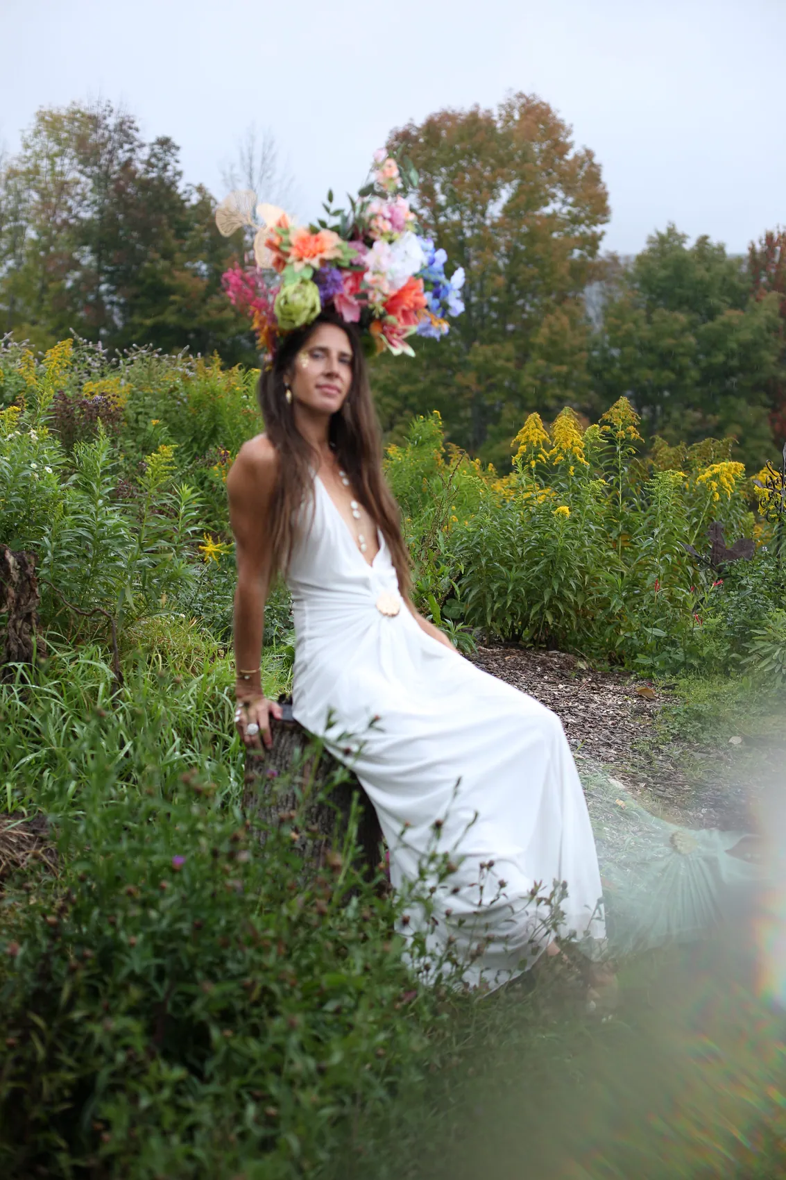 A woman in a long white gown seated in a wild garden, wearing a tall peach and pink crown