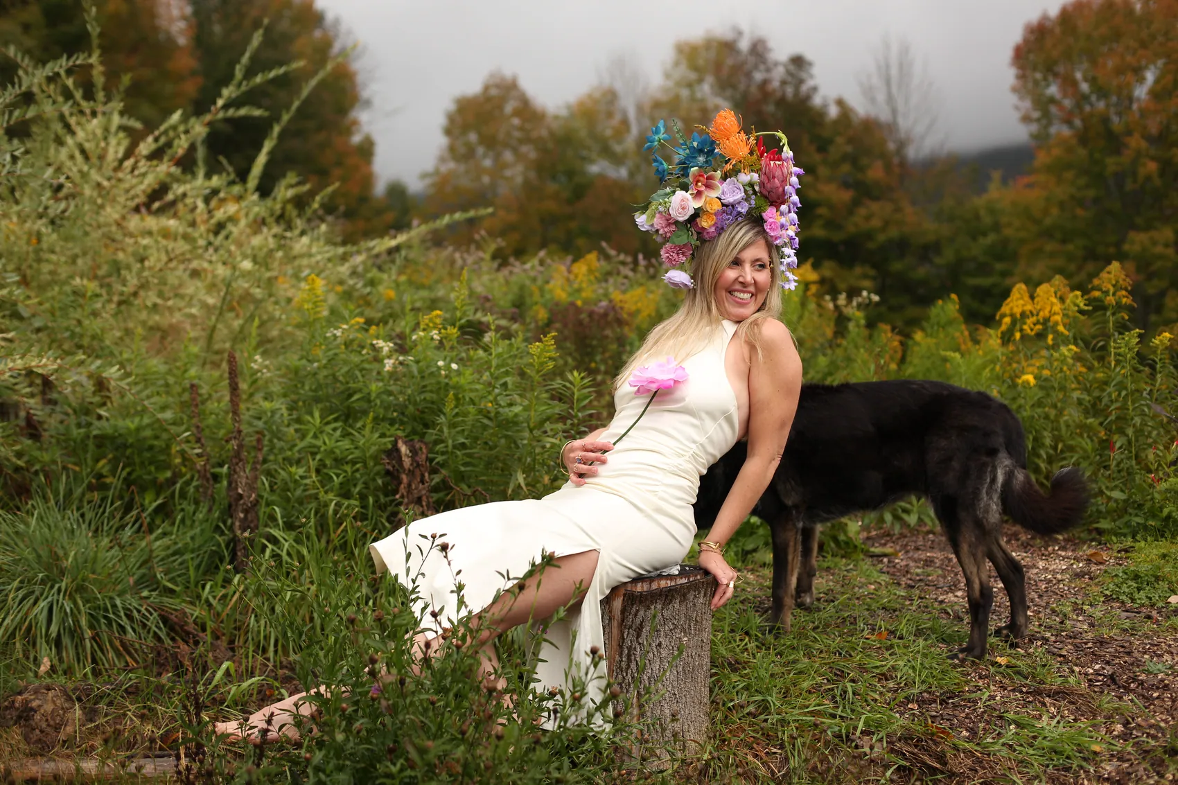A laughing woman in a cream gown seated beside a black dog, in a multicolour crown
