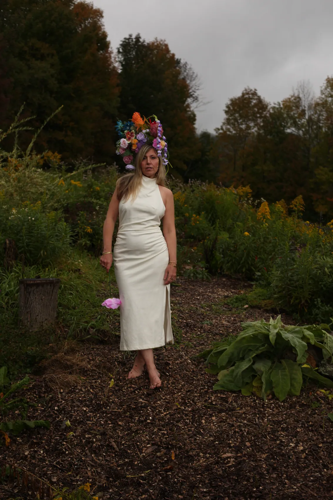 Daniela in a white halter column dress wearing a multicolour crown, full-length on a garden path