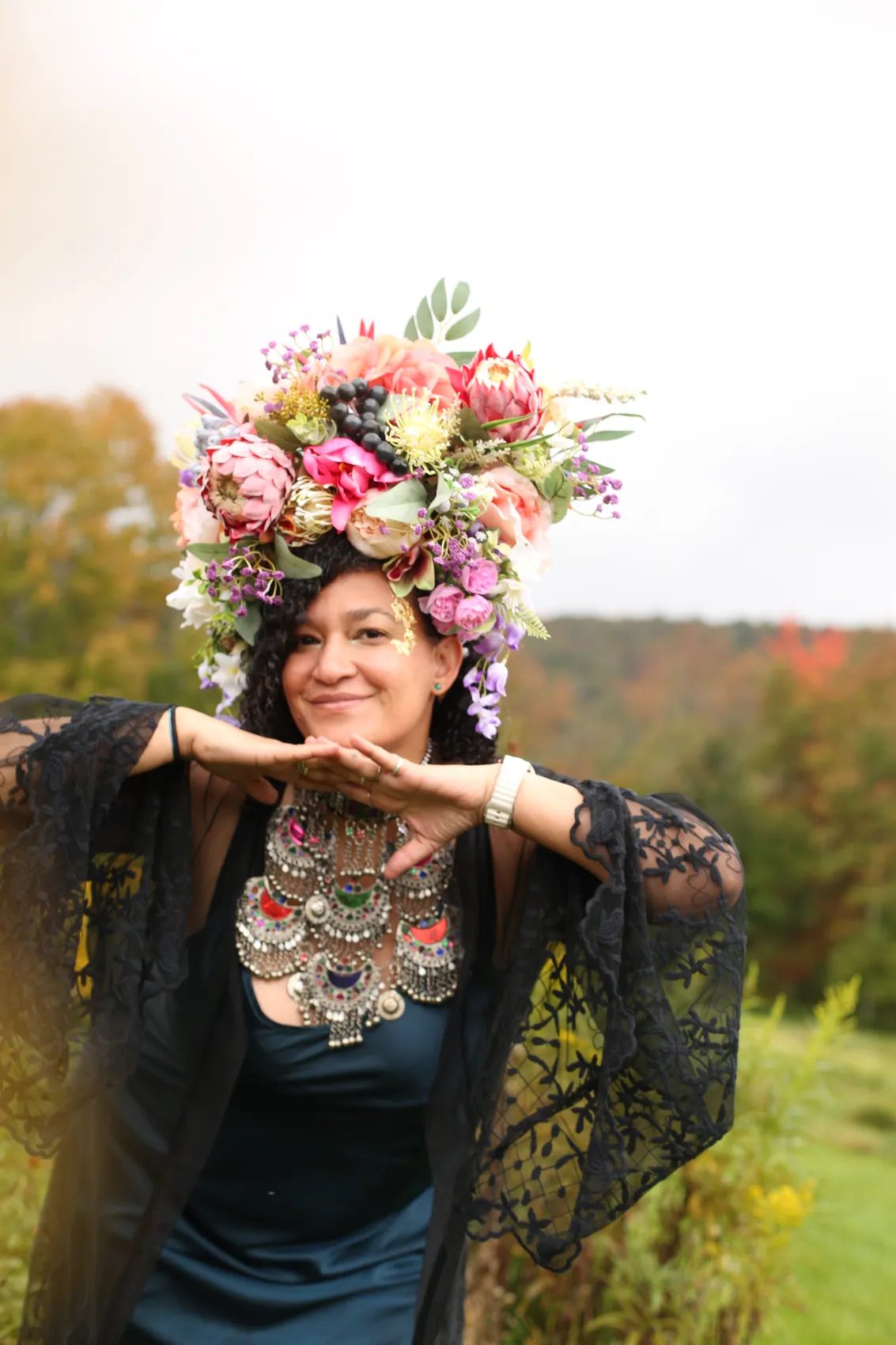 A woman in black lace and a statement necklace posing with her hands under her chin, in a crown of protea and peonies