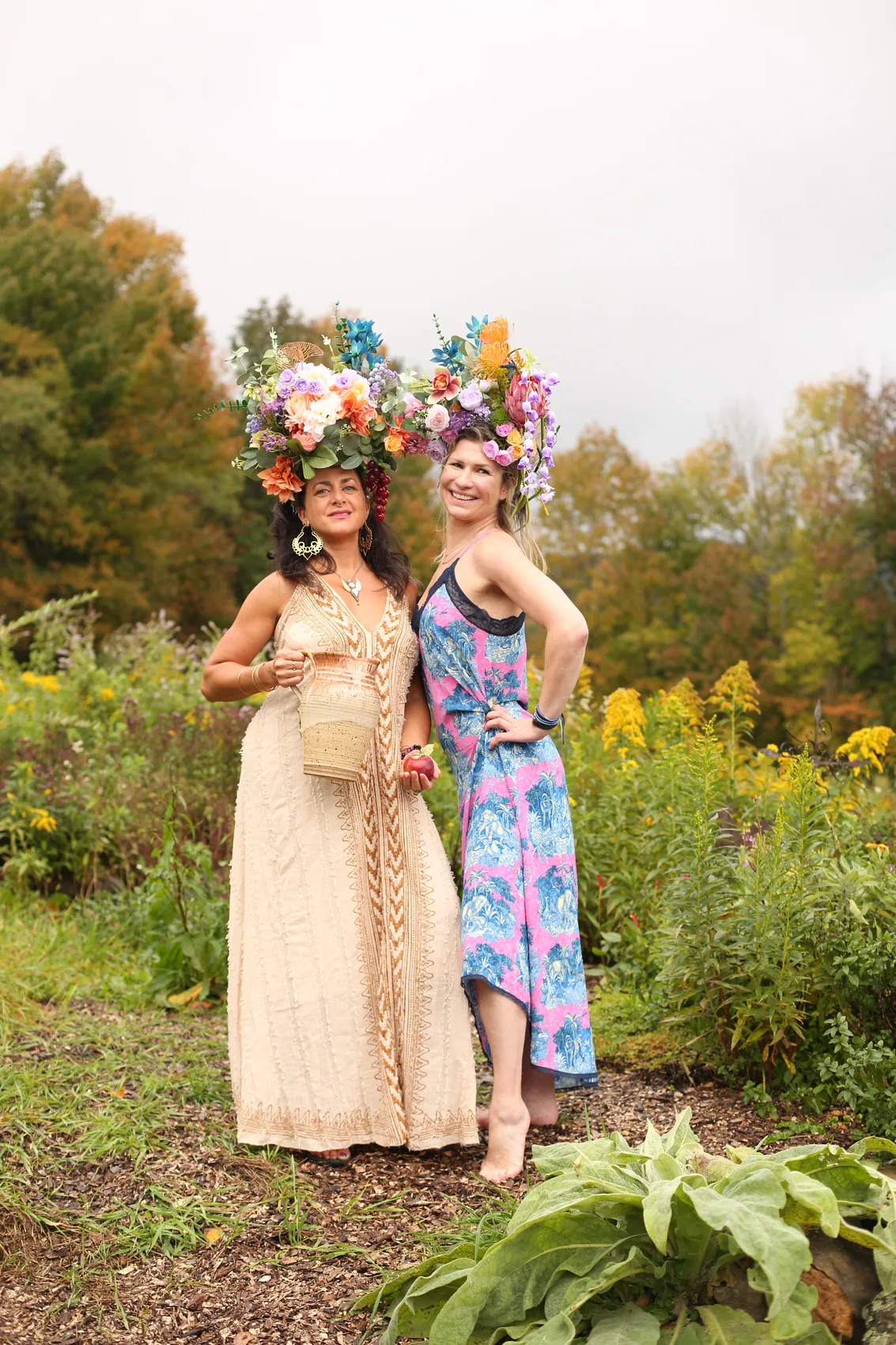 Two friends standing together, one in a cream gown and one in a blue and pink print dress, both in tall crowns