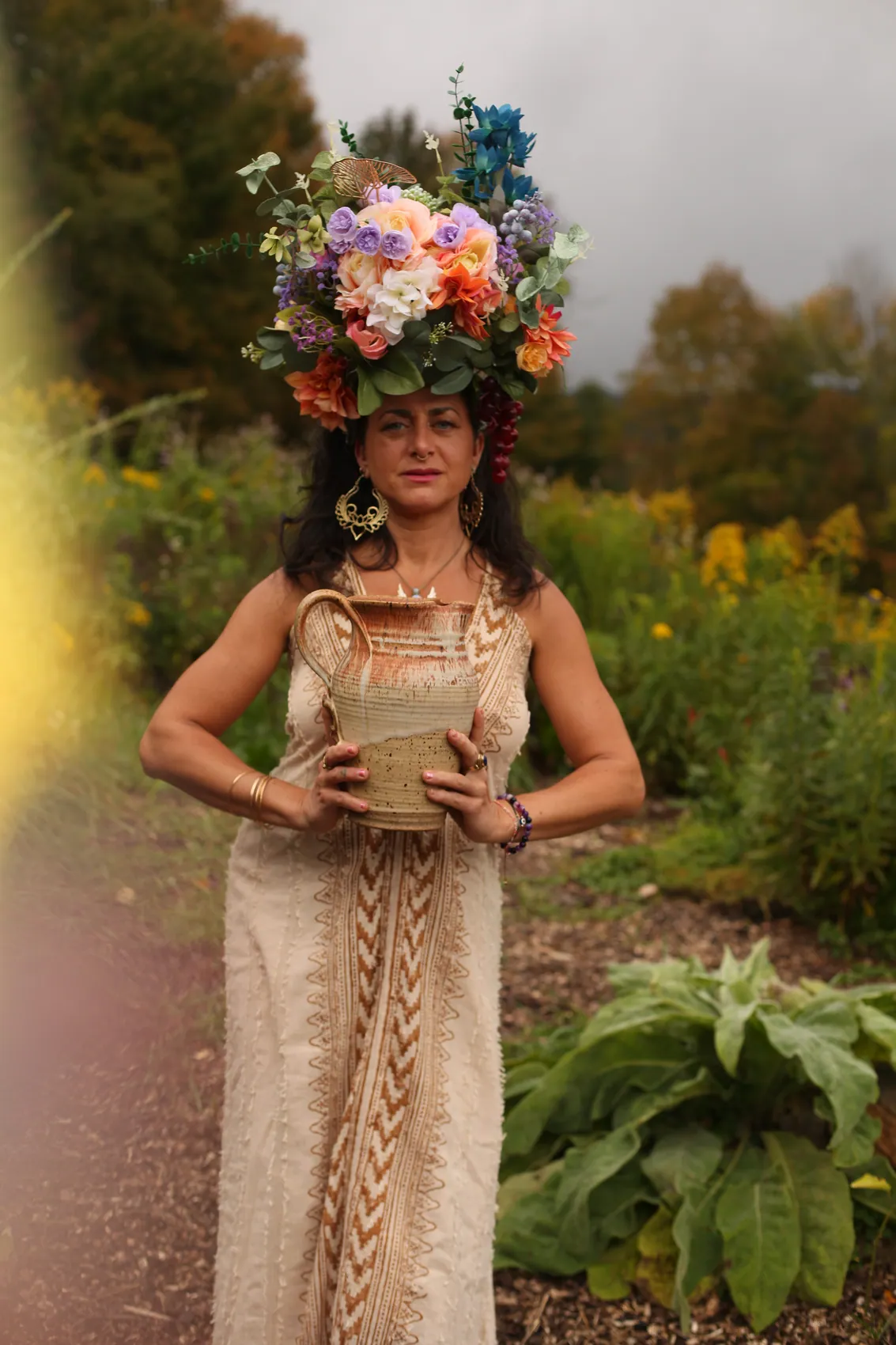 A woman in a cream knit gown holding a clay jug, wearing a tall crown of lilac and orange flowers