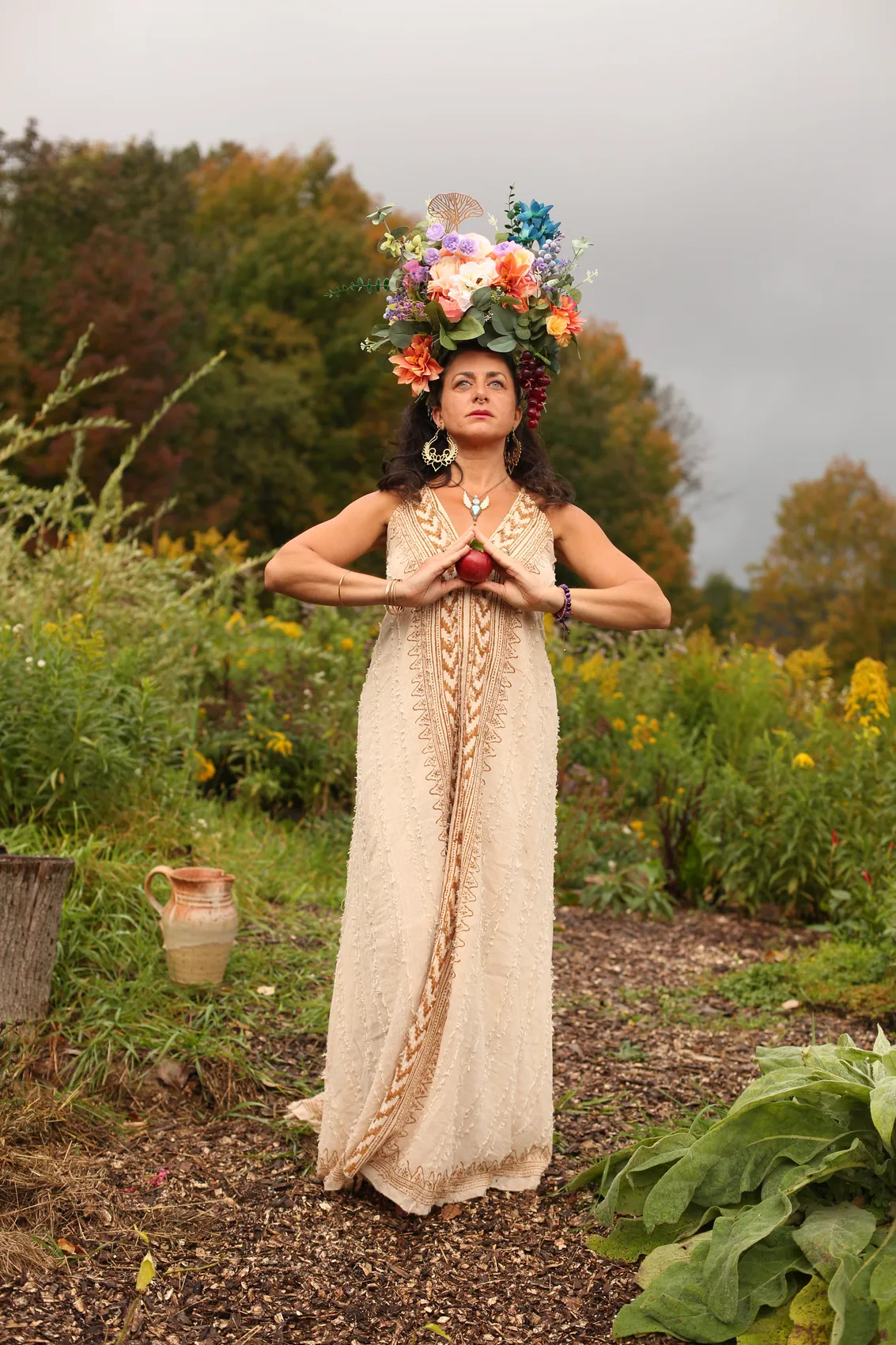 A woman in a cream gown and tall flower crown making a heart with her hands