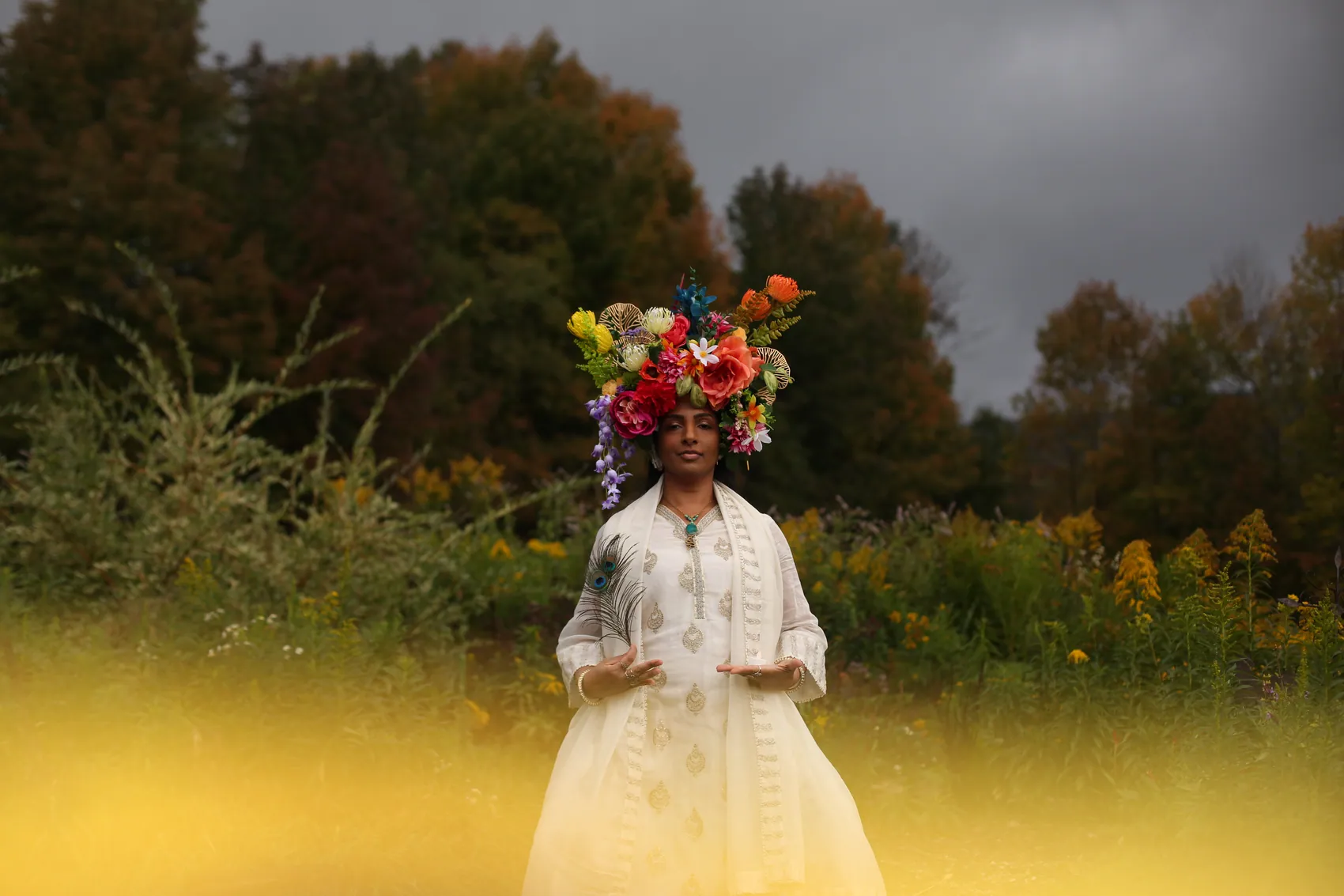 A woman in white embroidery wearing a crown of red, coral and yellow flowers under a stormy sky