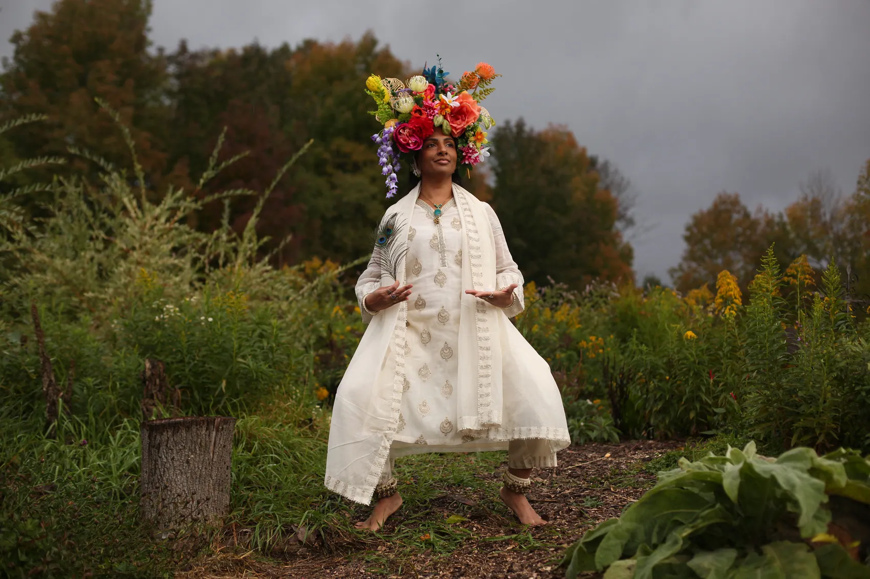 A woman in white embroidery in a grounded goddess pose, wearing a crown of red and coral flowers