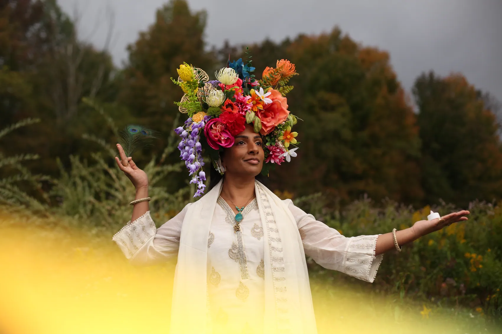 A woman in white embroidery with arms open wide, in a crown of red and coral flowers
