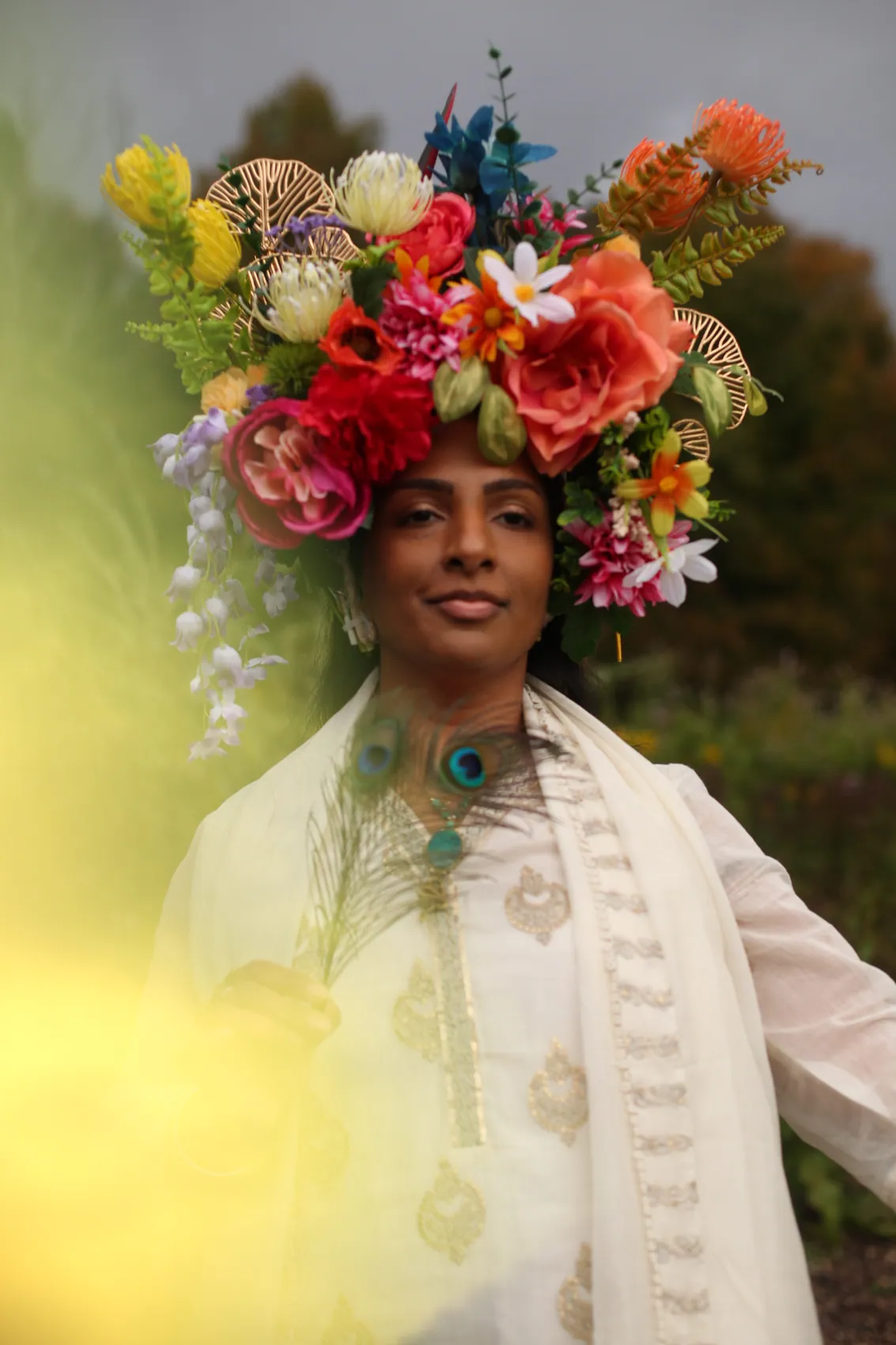 A woman in white embroidery wearing a tall crown of coral roses, protea and wisteria