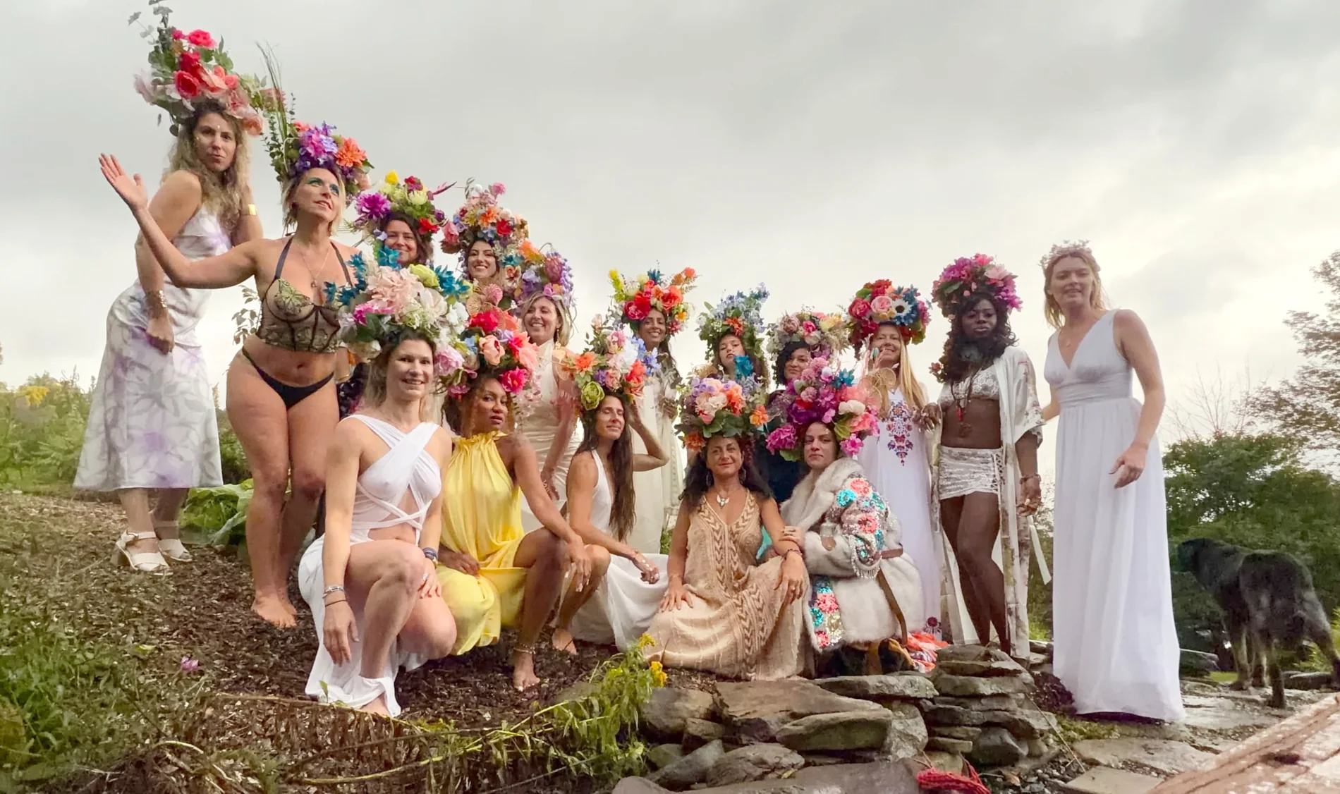 A circle of women in white and pastel dresses gathered on a hillside under a soft grey sky, each wearing a different handmade floral headdress
